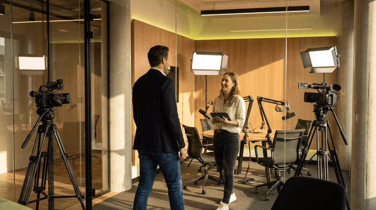 Homme en costume et femme à la tablette dans une salle vitrée équipée de deux caméras, de panneaux LED et de micros