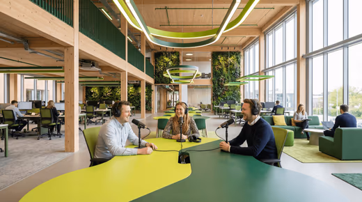 Trois collègues souriants enregistrent un podcast avec micros et casques au milieu d'un open space en bois végétalisé